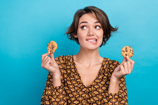 Photo of cute cheerful minded person biting lips arms hold cookies look empty space isolated on blue color background