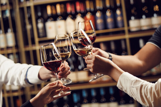 People in good mood clink glasses, celebrating together, photo focus on hands woman and man tasting degustation different types of wine, alcohol beverage to relax, bottles on shelves on background