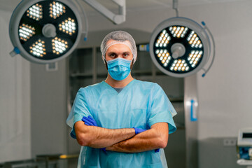 Fototapeta na wymiar Serious male surgeon in uniform and protective cap folds his hands in front of him and looks at the camera in operating room
