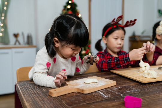 closeup of an adorable asian baby girl having fun carving her dough with a mold while making ginger cookies for Christmas at table with her sister