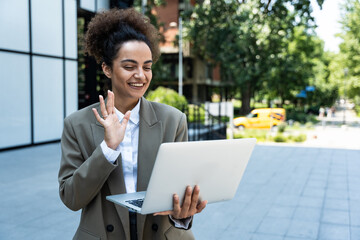 Portrait of confident successful young curly woman, formally dressed, business woman standing with laptop outdoors against the background of the business center, looks at camera, smiling