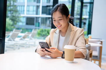 Cheerful Asian female freelancer enjoys chatting with co-workers on her smartphone while sitting at a table in a coffee shop.