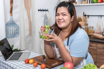 Asian fat woman standing in the kitchen, eating a salad, which she makes herself, which is a...