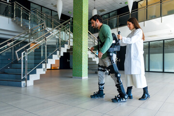 Mechanical exoskeleton, female doctor physiotherapist walking with disabled person helped by...