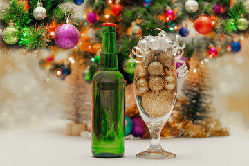 shiny christmas balls in a beer glass next to a green bottle and a decorated new year tree