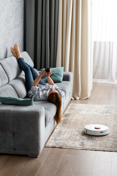 The girl lies on the sofa in the living room and controls a smart robot vacuum cleaner using a smartphone.
