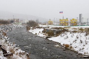 Magadanka River. Magadan, Magadan region, Russia. View of a playground and the Russian flag on the bank of a small river in a northern city. Cold snowy weather.