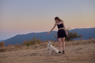 a beautiful, happy woman walks with a white dog in the mountains at sunset