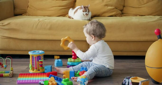 Blond toddler playing with builder constructor in living room. Boy child plays with toys sitting on floor of large apartment in morning