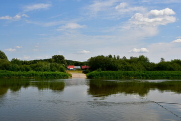 View of a massive lake, river, or pond surrounded from all sides with reeds, moss, forests, moors, and other flora spotted on a cloudy yet warm summer day next to some holiday resort located in Poland