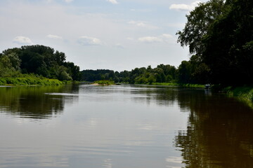 View of a massive lake, river, or pond surrounded from all sides with reeds, moss, forests, moors, and other flora spotted on a cloudy yet warm summer day next to some holiday resort located in Poland