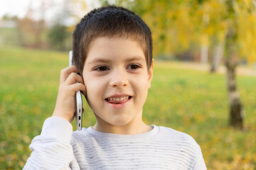 Handsome 6 year old boy talking on the phone while standing in park in autumn.