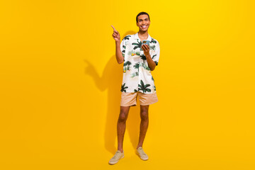Fototapeta na wymiar Full length photo of nice young male hold gadget point empty space dressed stylish palms print garment isolated on yellow color background