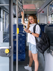 Naklejka premium Young urban woman stands inside the bus and uses her mobile phone. Using of mobile information devices in public transportation.