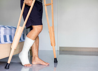 Close-up patient with broken leg in cast and bandage, man with leg splin is walking support with crutches at home
