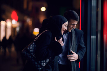 Fototapeta na wymiar Happy multicultural business couple walking together outdoors in an urban city street at night near a jewelry shopping store window.