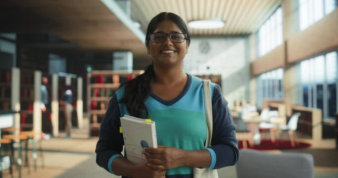 Portrait of a Cheerful Indian Student Standing in a Traditional Public Library. Young South Asian Female Looking at Camera and Smiling. Scholar Holding Academic STEM and Math Textbook