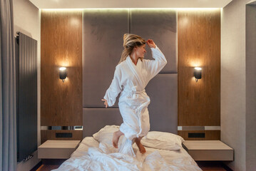 Woman having fun and jumping with bathrobe on bed in hotel room - obrazy, fototapety, plakaty