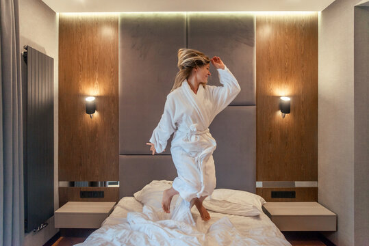 Woman having fun and jumping with bathrobe on bed in hotel room