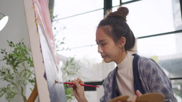 Young serious Asian woman Artist painting artwork at home workspace.