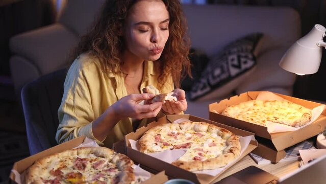 Excited curly haired young woman ordered many different pizzas to watch movie on laptop after hard work week at cozy home workplace Happy female enjoying watching favourite shows late at night 