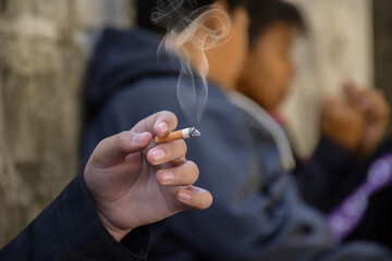 Closeup hand holding lighted cigarrette smoking of asian boys who starting to smoke in private area of school and house to avoid punishment from teachers or parents, new edited. - obrazy, fototapety, plakaty