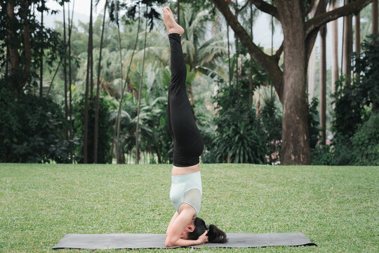 Asian woman doing yoga in Supported Headstand (Salamba Sirsasana) poses on mat in outdoor park.