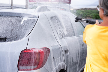 Back view of caucasian senior woman washing her car in a self-service car wash station using high...