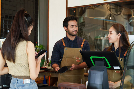 Barista employees coffee shop businessmen and small businesses Asian woman Standing cash register Asian female customer scans QR code tablet held by Indian man Behind cash register minimalist shop.