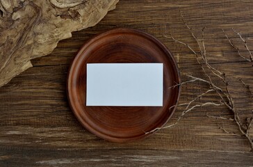 A mockup of a brown plate with white  card on dark old wooden table with dried plant branch and textured tree bark close-up.