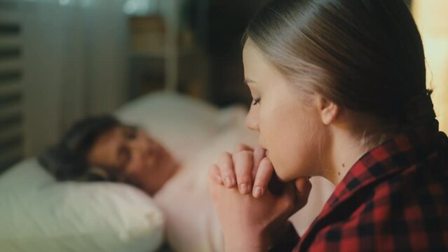 Sad granddaughter crying and praying near terminally ill granny in hospice ward