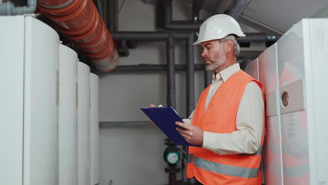 Grey bearded engineer in protective uniform making report about serviceability of power engines in control factory. Serious male with clipboard standing and showing professionalism in industry.