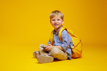 Side view of excited pupil kid wearing blue shirt and yellow backpack, holding a phone in hands while sitting on studio floor with crossed legs, isolated over yellow background.