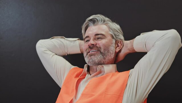 Satisfied caucasian old man wearing orange vest sitting on chair while holding clenched hands behind head. Tired inspector resting at workplace during lunch break. Isolated on black background.