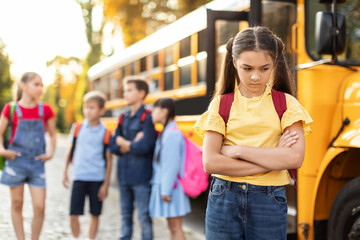 Unhappy young girl standing by herself next to yellow school bus - obrazy, fototapety, plakaty