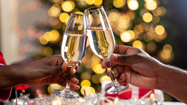 Close em mãos de duas pessoas fazendo um brinde com espumante em taças e luzes de Natal ao fundo.