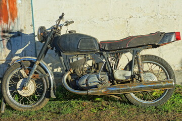 one old  iron classical  heavy big comfortable black  broken    dirty made in the ussr retro  motorcycle stands  near the white wall  on the ground in the street 
