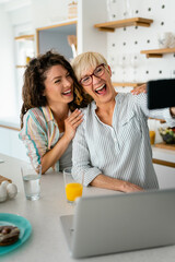 Funny senior mother with eyeglasses and adult smiling daughter taking selfies in the kitchen.