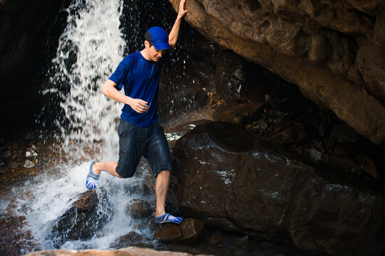 A young man in a blue t-shirt and hat walks under a waterfall in a sandstone alcove, closeup