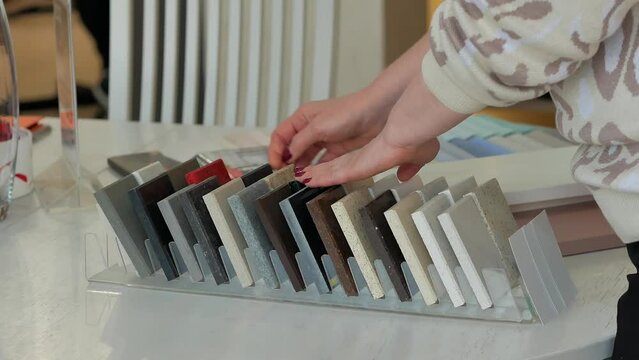 A woman with a beautiful manicure is sorting through samples of stone countertops in a furniture store on a white wooden table. Purchase of kitchen furniture by individual design, choice of material