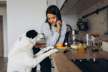 Beautiful caucasian lady with her black and white big dog are preparing autumn winter sea buckthorn tea with orange, cinnamon and honey. Healthy drinks concept - obrazy, fototapety, plakaty