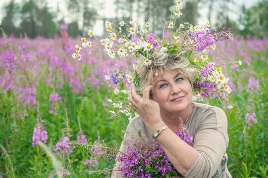 Beautiful woman 60 years old in nature with a wreath of flowers in her hair. Beautiful elderly happy woman smiling. Field and wreath of flowers of cypress Chamaenerion angustifolium