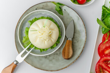 Sliced Italian cheese burrata is served on a round plate with basil and tomatoes with a fork and knife on the background of an old tree. Overhead view