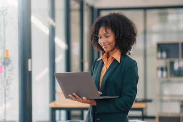 Young african american happy businesswoman standing and working on laptop by window in office.