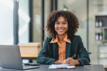 Young african american businesswoman using laptop computer, taking notes, working project in modern office. Financier working with documents at workplace.