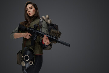 Middle Eastern-looking woman dressed in survivalist clothing posing with a rifle against a gray background, portraying strength and resilience