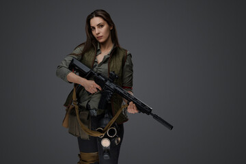 Naklejka premium Middle Eastern-looking woman dressed in survivalist clothing posing with a rifle against a gray background, portraying strength and resilience