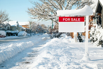 Home for sale sign with winter background scene on a sunny day. Red for sale realtor signage on sign post defocused snowed cars, homes and  street. Real estate and working in winter. Selective focus.