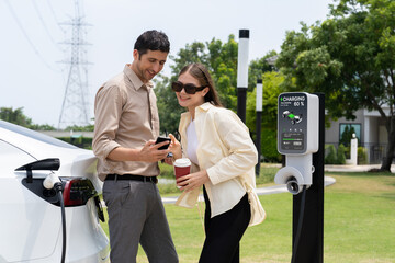 Young couple with coffee pay for electricity while recharge EV car battery at charging station connected to electrical power grid tower as electric industrial for eco friendly car travel. Expedient