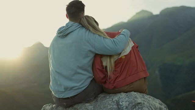 Hiking, view of mountain and happy couple hugging, relax on outdoor adventure and freedom in nature from back. Trekking, rock climbing and love, man and woman at sunset sky sitting on cliff together.